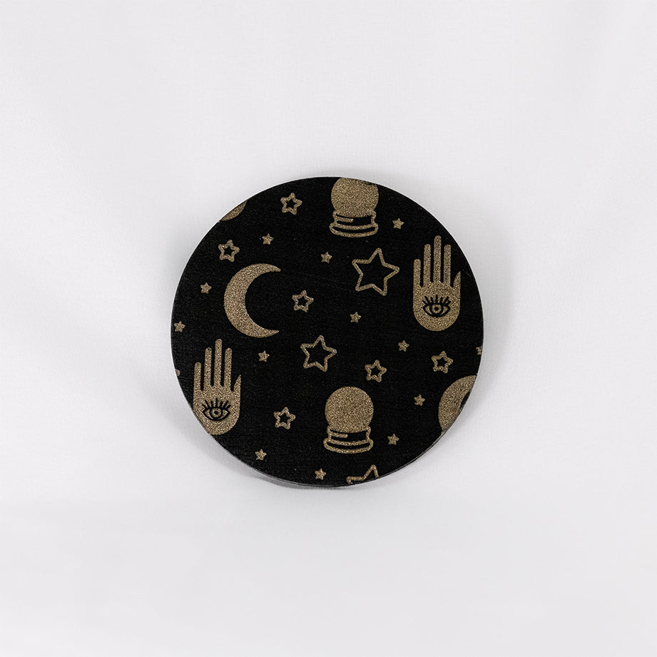 A round black coaster engraved with gold stars, crescent moons, a palm with an eye in it, and crystal balls. The coaster is on a white background.