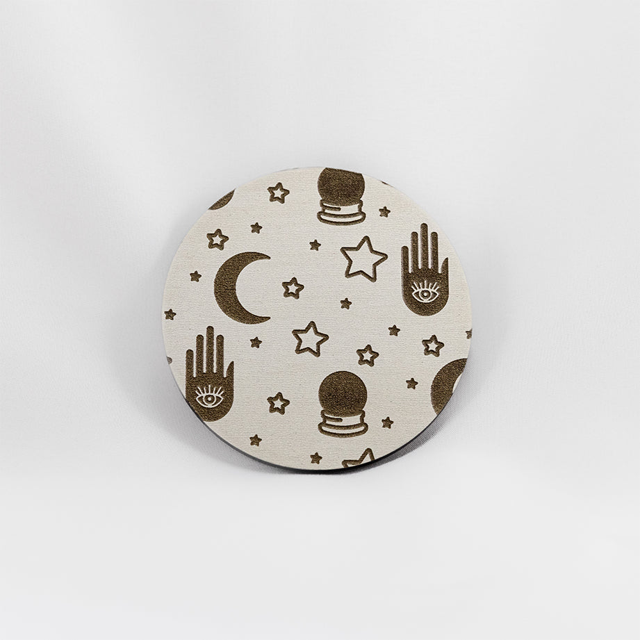 A round white coaster engraved with gold stars, crescent moons, a palm with an eye in it, and crystal balls. The coaster is on a white background.
