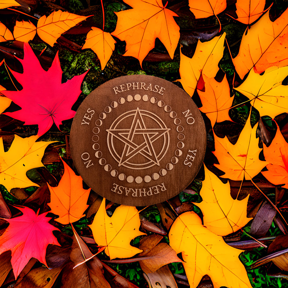 Circular Baltic birch laser-cut pendulum board engraved around its border with the words "yes", "no", "rephrase", the board has a pentacle in the centre encircled with the different moon phases and lays on the leaf covered ground.