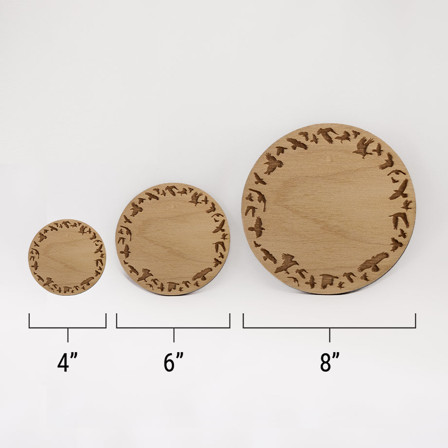 Three different sizes of the circular wooden laser-cut crystal grid engraved a border of ravens in flight against a white background with a ruler underneath all three. The smallest measuring 4", the middle 6", and the largest 8".