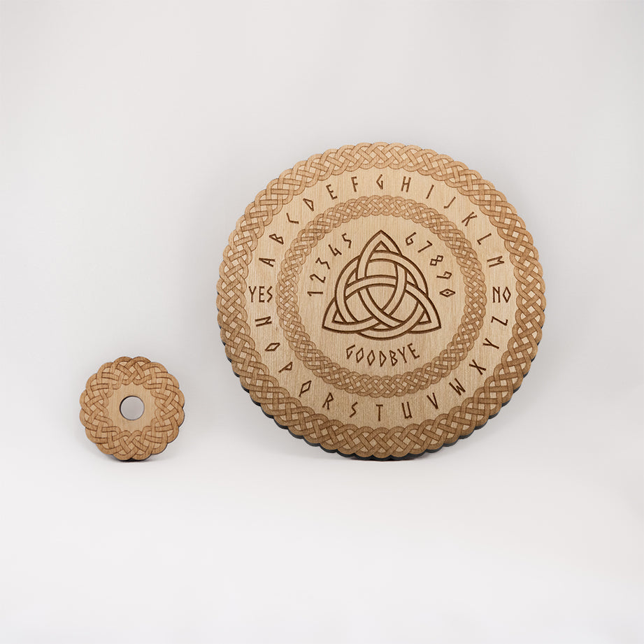 Circular Baltic birch laser-cut spirit board engraved with the words "yes", "no", "goodbye", as well as the entire alphabet. The board has the Celtic Triquetra symbol in the centre and a decorative die-cut braid border. To the side of the spirit board there is a small circular planchette with a matching die-cut braid border.