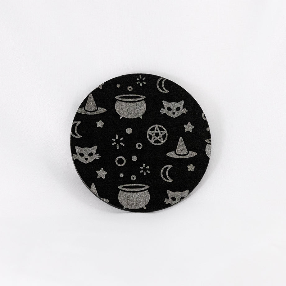 A round black coaster engraved with silver witchy symbols, including a bubbling cauldron, a pointed witch's hat, a crescent moon, a black cat, and a pentacle. The coaster is on a white background.