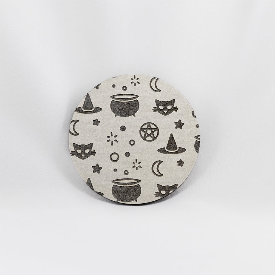A round white coaster engraved with silver witchy symbols, including a bubbling cauldron, a pointed witch's hat, a crescent moon, a black cat, and a pentacle. The coaster is on a white background.