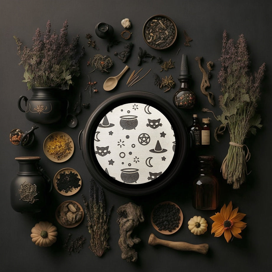 A round white coaster engraved with silver witchy symbols, including a bubbling cauldron, a pointed witch's hat, a crescent moon, a black cat, and a pentacle. The coaster is in a black cauldron, surrounded by small pumpkins, dried flowers, small dishes with dried herbs, and small potion bottles.