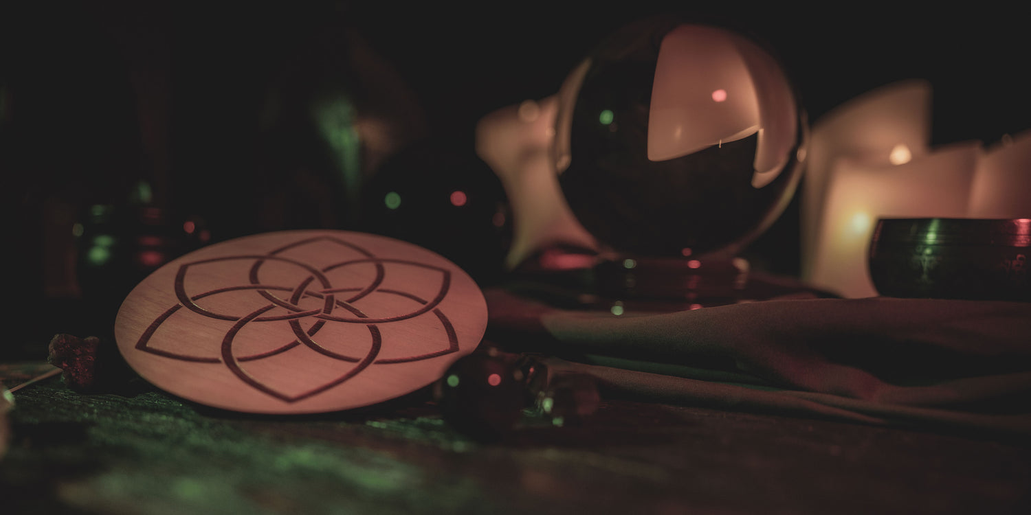 A small round wooden crystal grid with a celtic knot engraved on it sits on a table with a crystal ball and miscellaneous crystals in the background