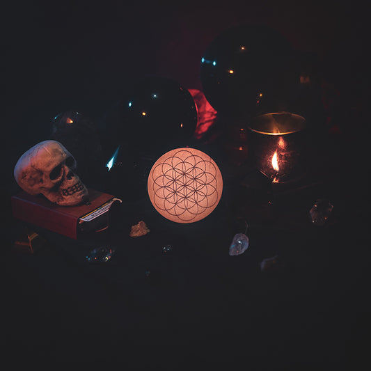 A circular wooden laser-cut crystal grid engraved with the Flower of Life. The grid sits amongst candles, crystal balls, a skull, tarot cards and assorted crystals.