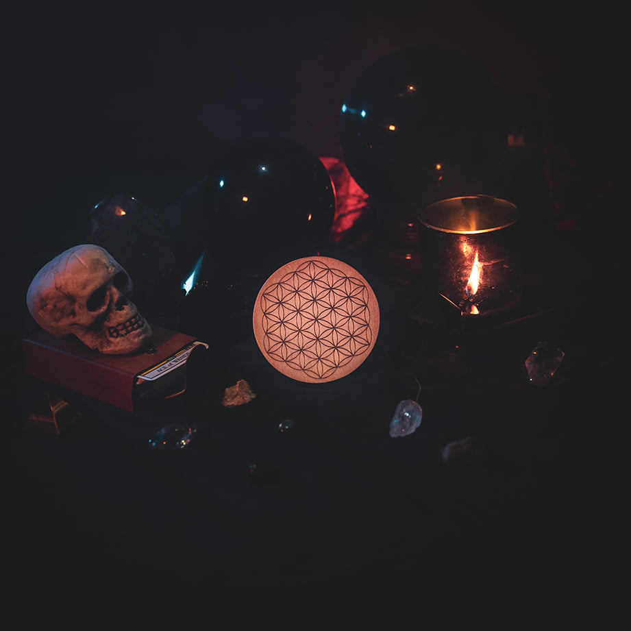 A circular wooden laser-cut crystal grid engraved with the Flower of Life. The grid sits amongst candles, crystal balls, a skull, tarot cards and assorted crystals.