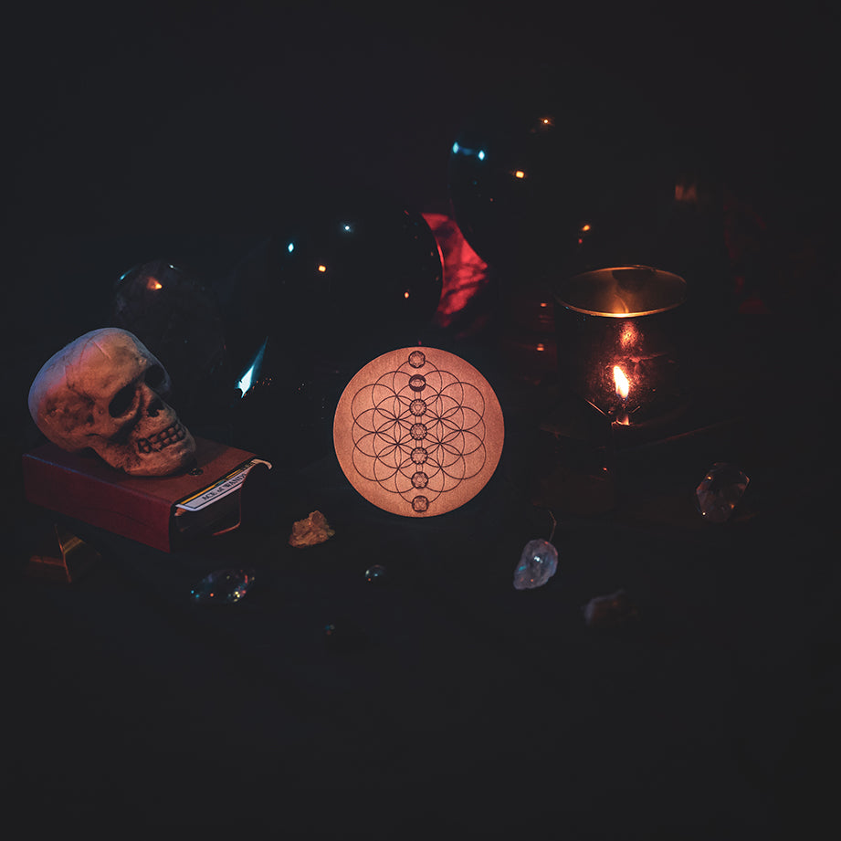 A circular wooden laser-cut crystal grid engraved with the Flower of Life and the seven Chakras. The grid sits amongst candles, crystal balls, a skull, tarot cards and assorted crystals.