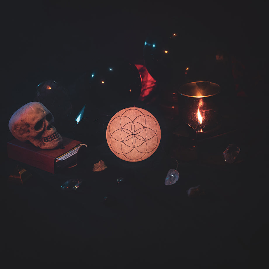 A circular wooden laser-cut crystal grid engraved with the Seed of Life. The grid sits amongst candles, crystal balls, a skull, tarot cards and assorted crystals.