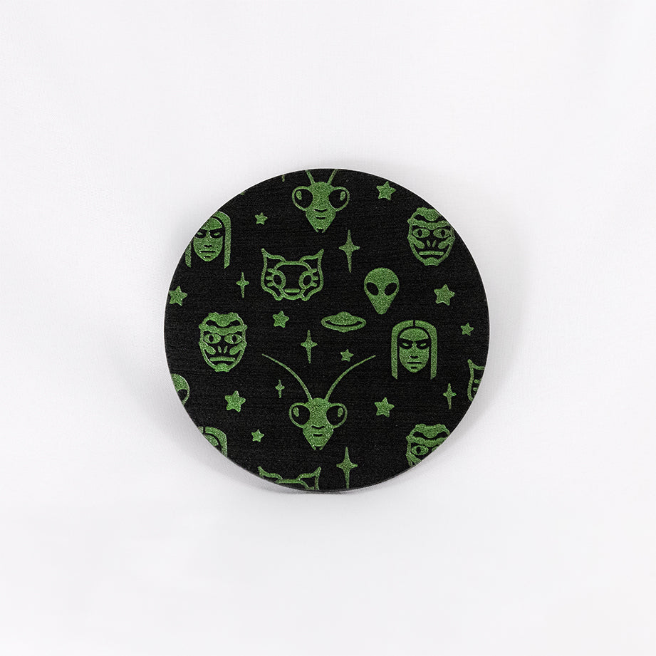 A round black coaster engraved with green aliens and space themed items; grey aliens, nordic aliens, mantis aliens, goblin aliens, stars and a flying saucer. The coaster is on a white background.