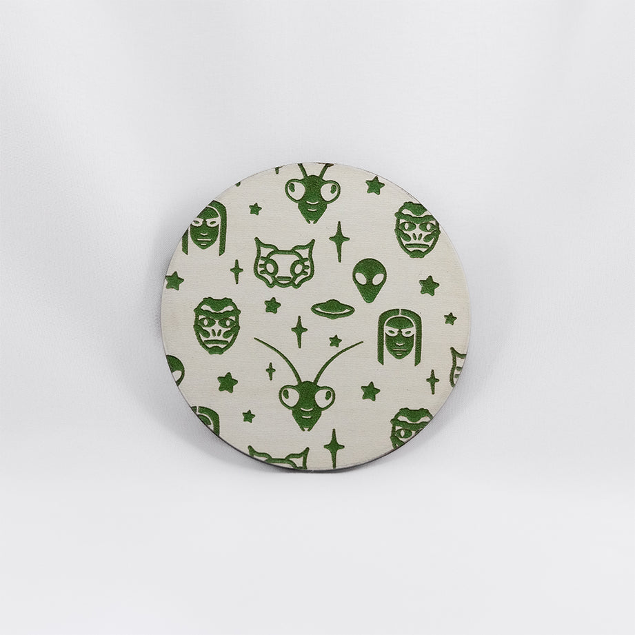A round white coaster engraved with green aliens and space themed items; grey aliens, nordic aliens, mantis aliens, goblin aliens, stars and a flying saucer. The coaster is on a white background.
