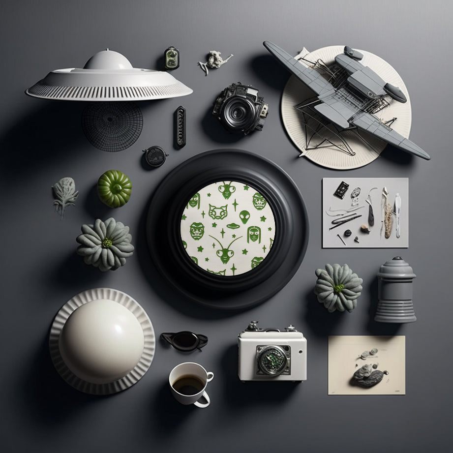 A round white coaster engraved with green aliens and space themed items; grey aliens, nordic aliens, mantis aliens, goblin aliens, stars and a flying saucer. The coaster is on a grey dish framed by flying saucer toys, strange grey gourds, and small metallic tools. 