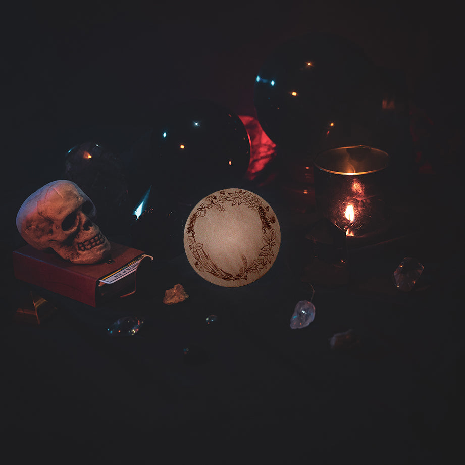 A circular wooden laser-cut crystal grid engraved with a border made up of Apple, Yew, Hazel, Edler, Elm, Willow, Oak, Alder, and Ash tree branches. The grid sits amongst candles, crystal balls, a skull, tarot cards and assorted crystals.