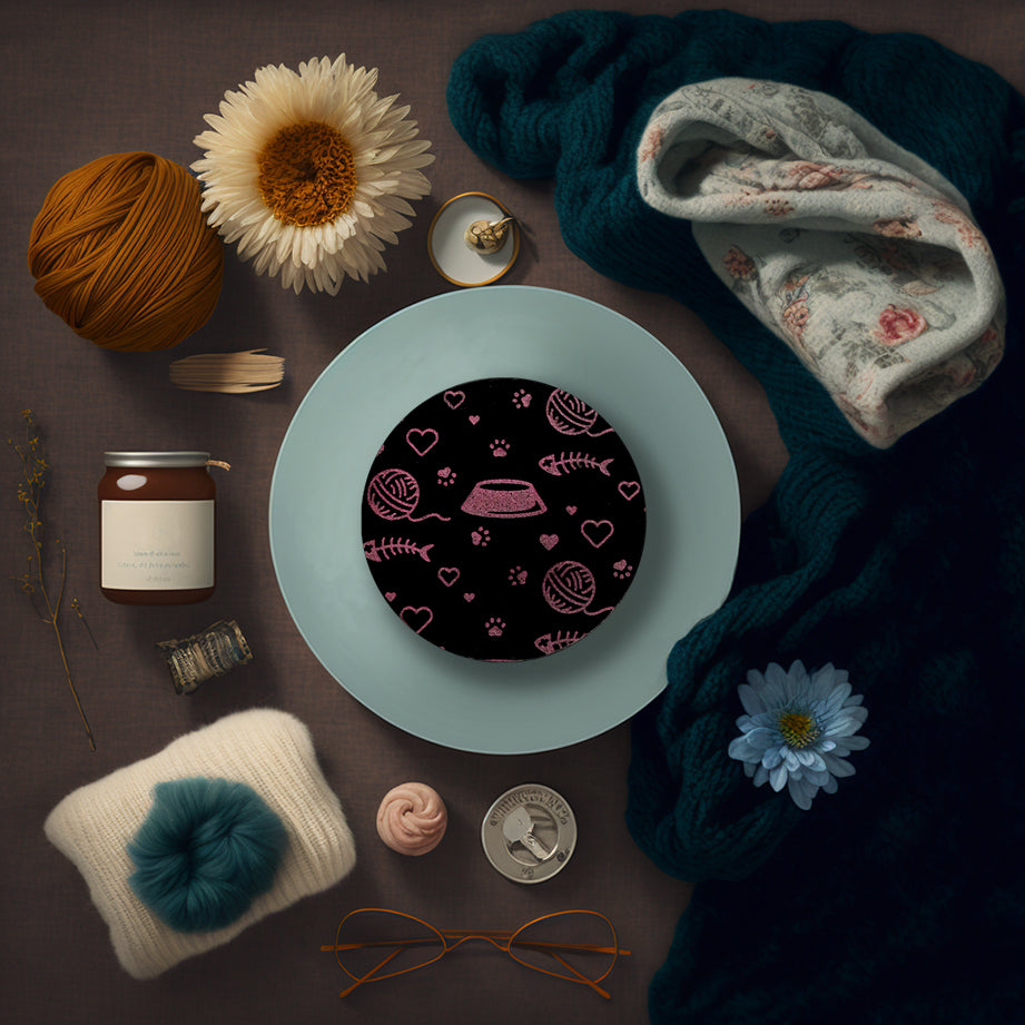 A round black coaster engraved with pink cat themed items; paw prints, a ball of yarn, hearts, a kibble dish, and a fish skeleton. The coaster is on a blue dish framed by a blanket, yarn, flowers, cup of tea, and glasses.