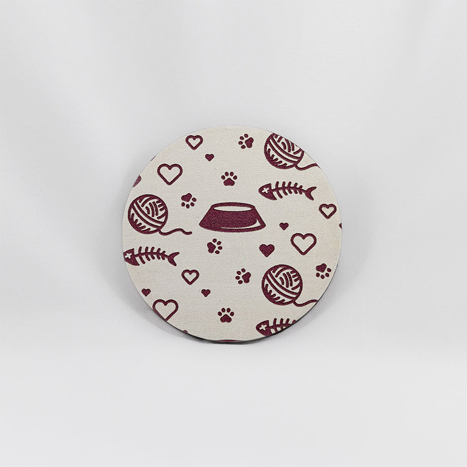 A round white coaster engraved with pink cat themed items; paw prints, hearts, a kibble dish, and a fish skeleton. The coaster is on a white background.