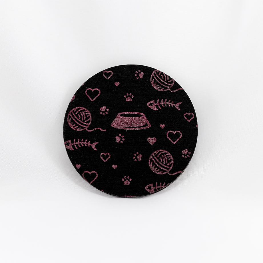 A round black coaster engraved with pink cat themed items; paw prints, hearts, a kibble dish, and a fish skeleton. The coaster is on a white background.
