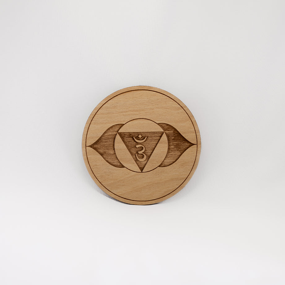 A circular wooden laser-cut crystal grid engraved with the third eye chakra symbol with a plain white background. 