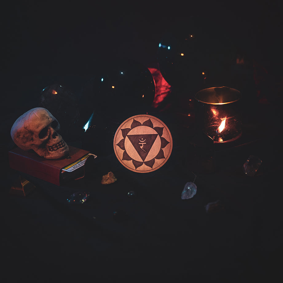 A circular wooden laser-cut crystal grid engraved with the solar plexus chakra symbol. The grid sits amongst candles, crystal balls, a skull, tarot cards and assorted crystals.