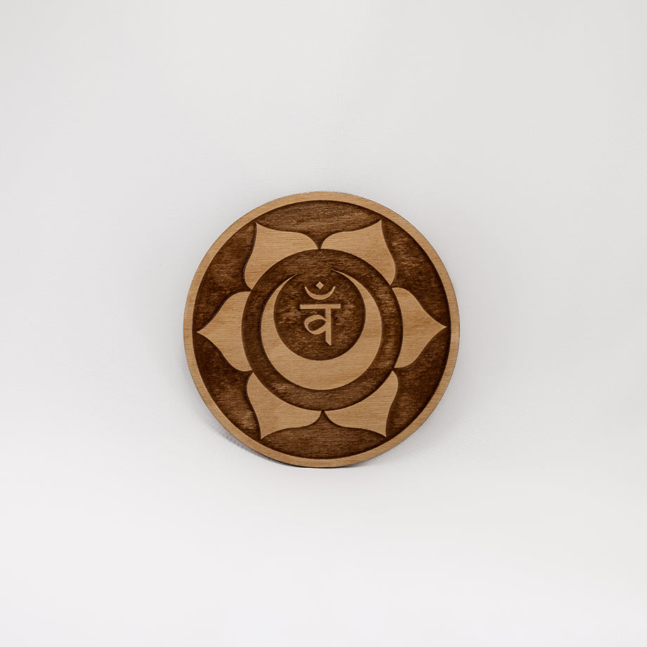 A circular wooden laser-cut crystal grid engraved with the sacral chakra symbol with a plain white background. 