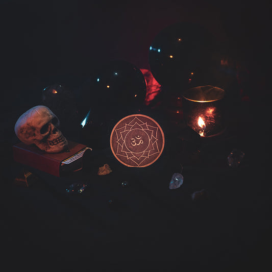 A circular wooden laser-cut crystal grid engraved with the crown chakra symbol. The grid sits amongst candles, crystal balls, a skull, tarot cards and assorted crystals.