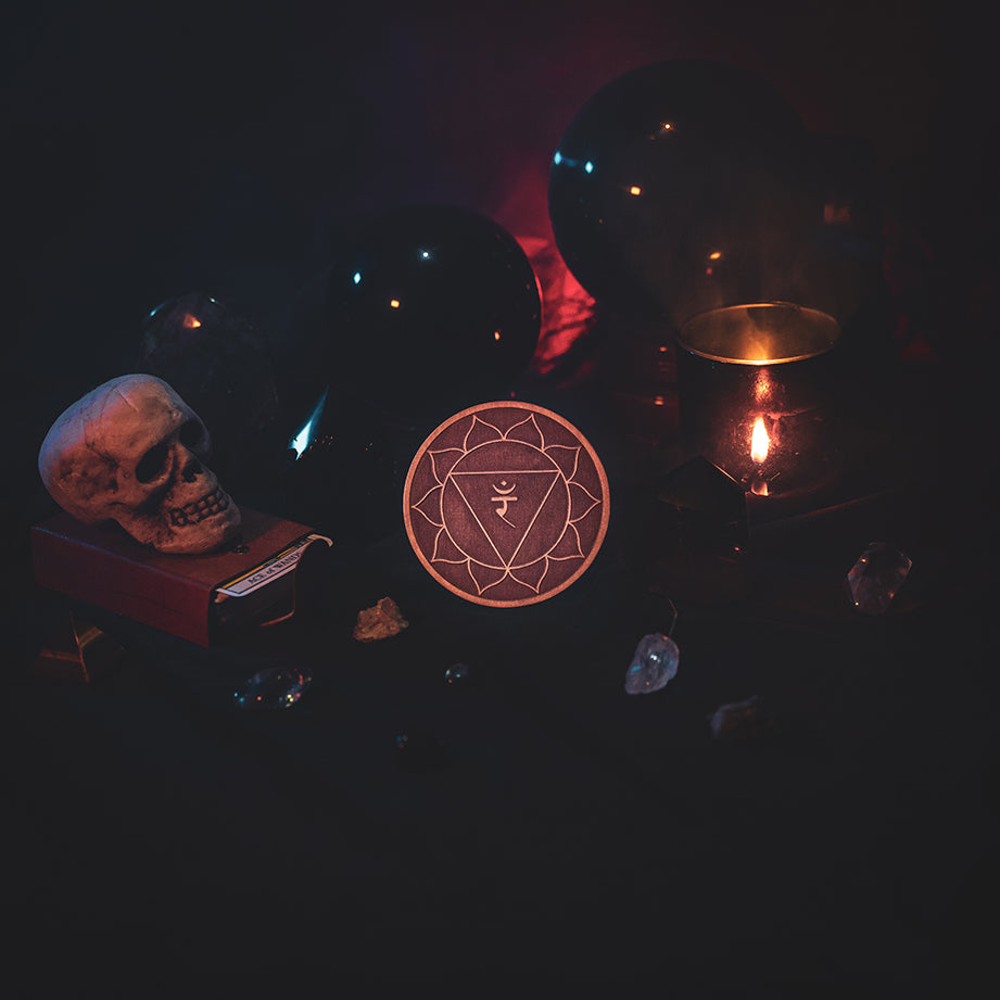 A circular wooden laser-cut crystal grid engraved with the solar plexus chakra symbol. The grid sits amongst candles, crystal balls, a skull, tarot cards and assorted crystals.