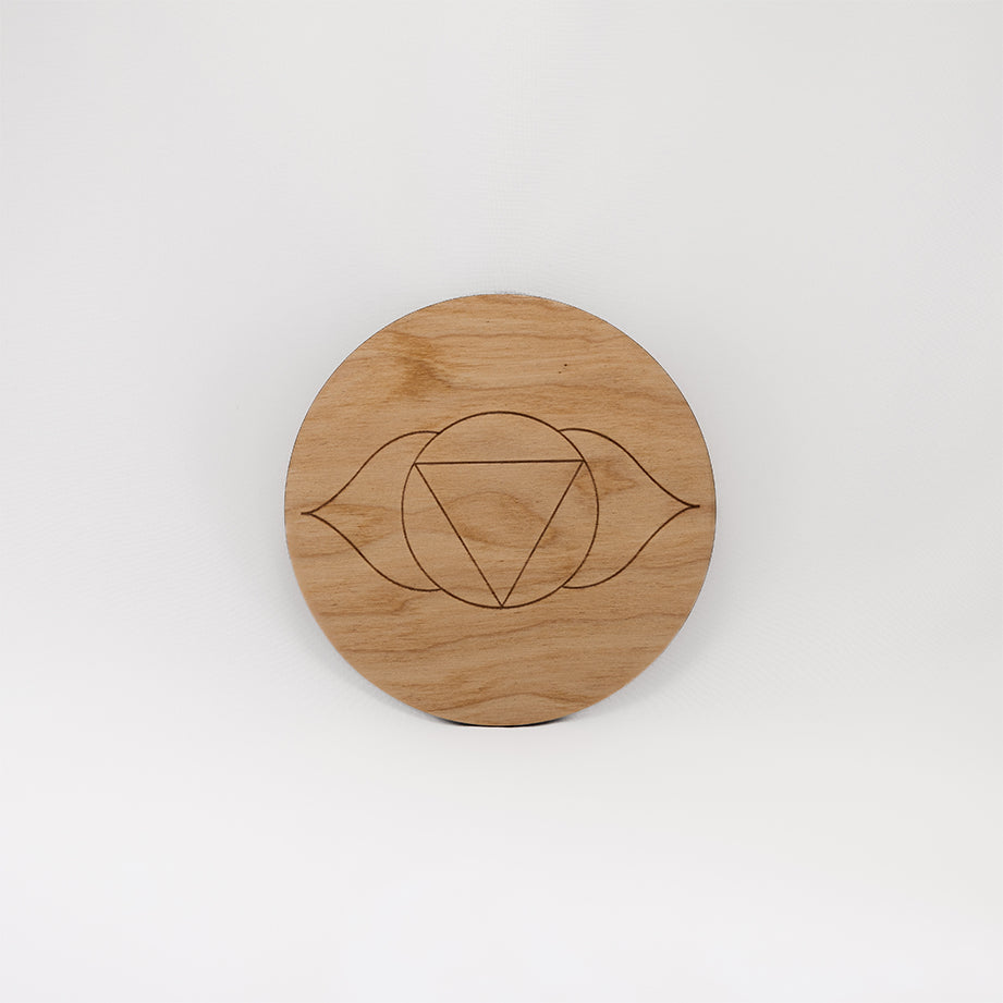 A circular wooden laser-cut crystal grid engraved with the third eye chakra symbol with a plain white background. 