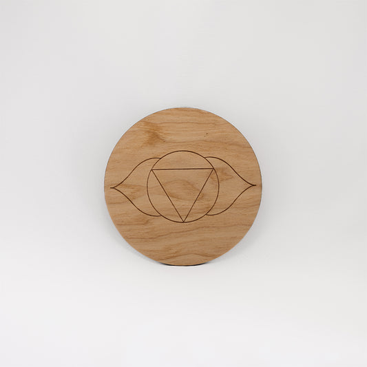 A circular wooden laser-cut crystal grid engraved with the third eye chakra symbol with a plain white background. 