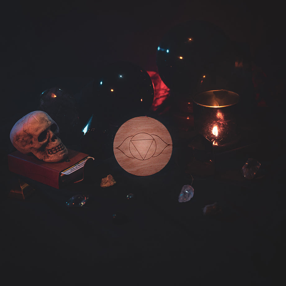 A circular wooden laser-cut crystal grid engraved with the third eye chakra symbol. The grid sits amongst candles, crystal balls, a skull, tarot cards and assorted crystals.