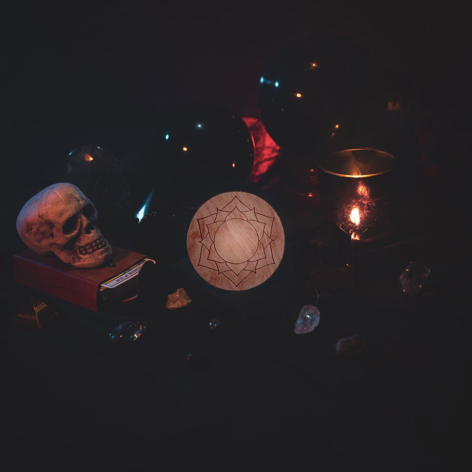 A circular wooden laser-cut crystal grid engraved with the crown chakra symbol. The grid sits amongst candles, crystal balls, a skull, tarot cards and assorted crystals.