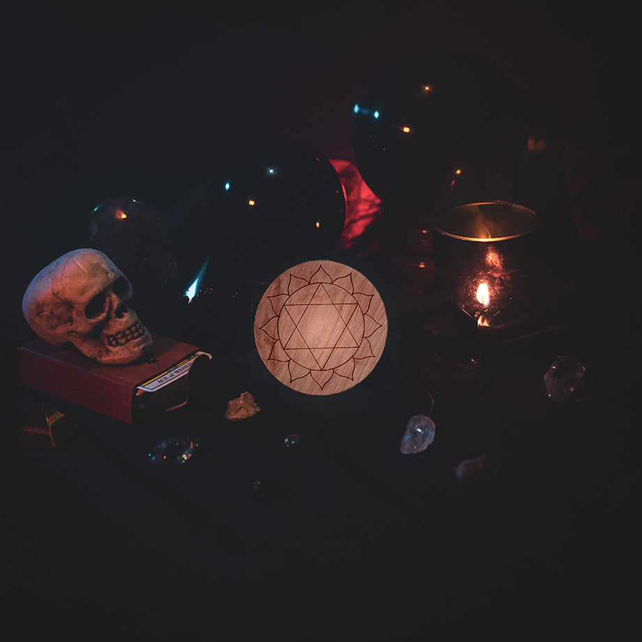 A circular wooden laser-cut crystal grid engraved with the third heart chakra symbol. The grid sits amongst candles, crystal balls, a skull, tarot cards and assorted crystals.