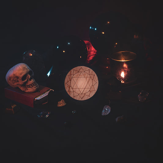 A circular wooden laser-cut crystal grid engraved with the third heart chakra symbol. The grid sits amongst candles, crystal balls, a skull, tarot cards and assorted crystals.