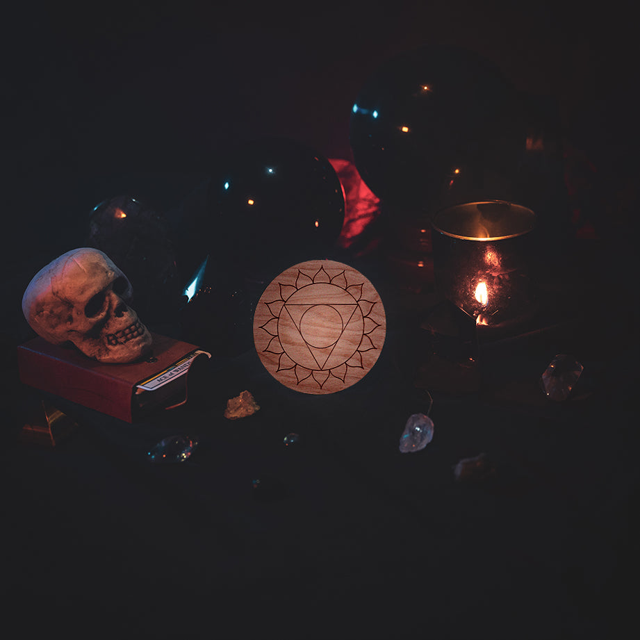 A circular wooden laser-cut crystal grid engraved with the throat chakra symbol. The grid sits amongst candles, crystal balls, a skull, tarot cards and assorted crystals.