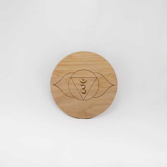 A circular wooden laser-cut crystal grid engraved with the third eye chakra symbol with a plain white background. 