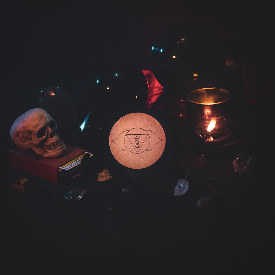 A circular wooden laser-cut crystal grid engraved with the third eye chakra symbol. The grid sits amongst candles, crystal balls, a skull, tarot cards and assorted crystals.