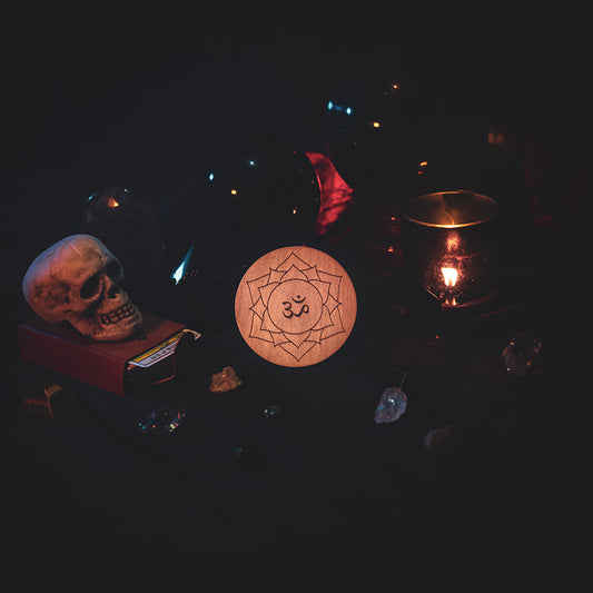 A circular wooden laser-cut crystal grid engraved with the crown chakra symbol. The grid sits amongst candles, crystal balls, a skull, tarot cards and assorted crystals.