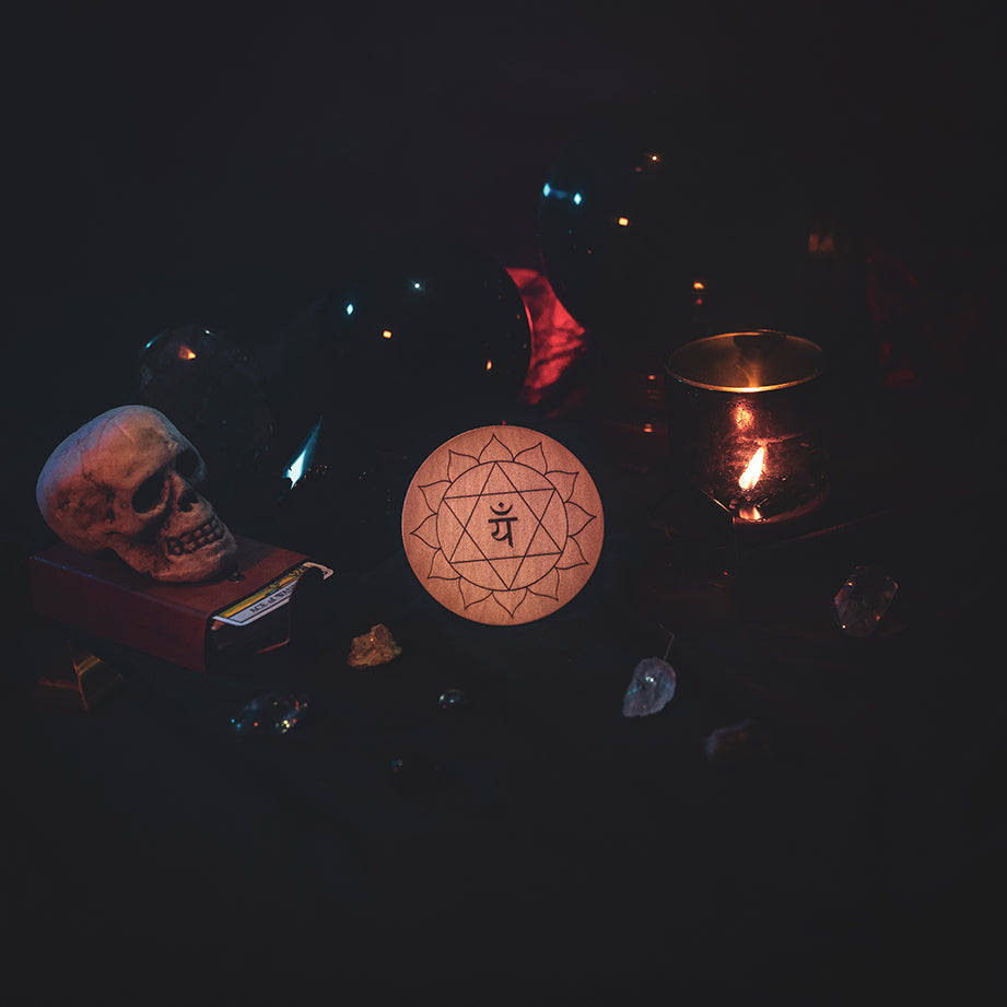 A circular wooden laser-cut crystal grid engraved with the third heart chakra symbol. The grid sits amongst candles, crystal balls, a skull, tarot cards and assorted crystals.