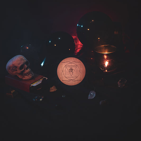 A circular wooden laser-cut crystal grid engraved with the root chakra symbol. The grid sits amongst candles, crystal balls, a skull, tarot cards and assorted crystals.