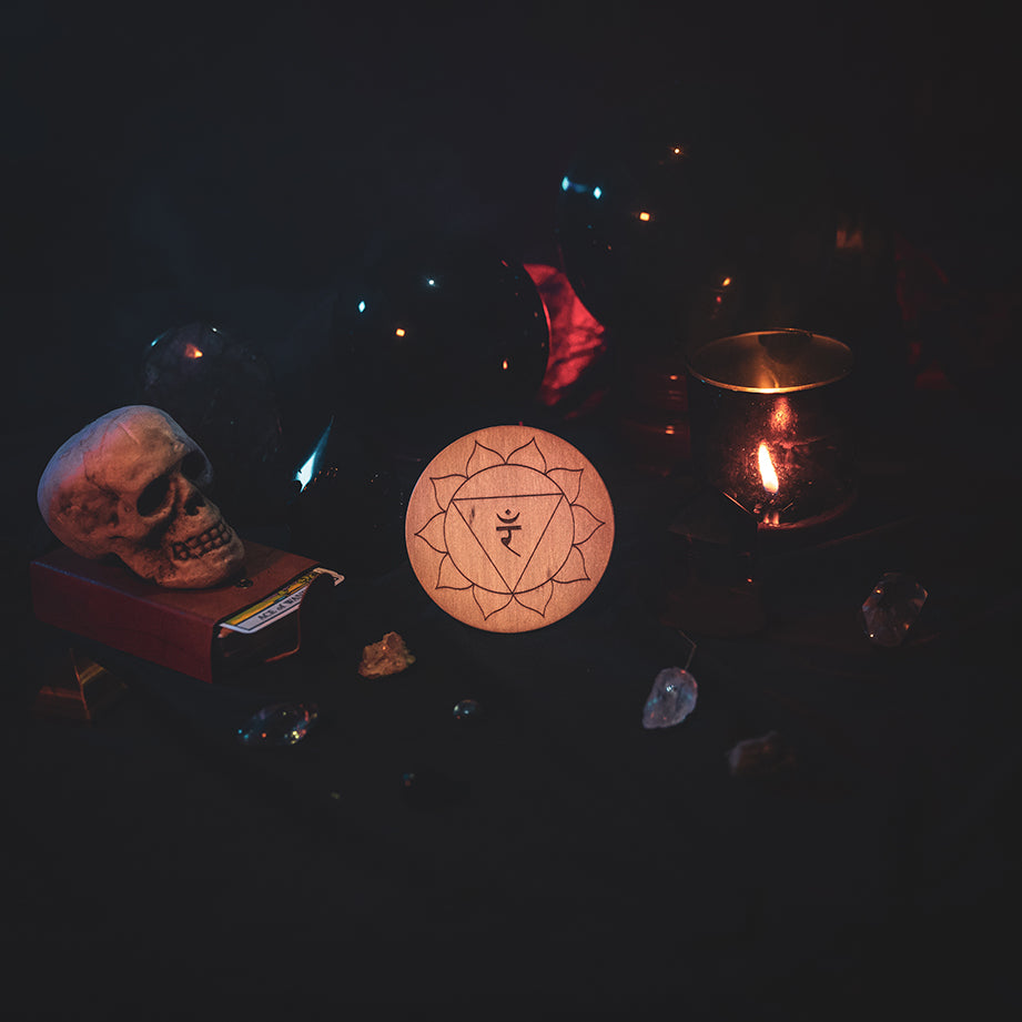 A circular wooden laser-cut crystal grid engraved with the solar plexus chakra symbol. The grid sits amongst candles, crystal balls, a skull, tarot cards and assorted crystals.