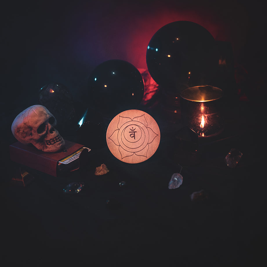 A circular wooden laser-cut crystal grid engraved with the sacral chakra symbol. The grid sits amongst candles, crystal balls, a skull, tarot cards and assorted crystals.