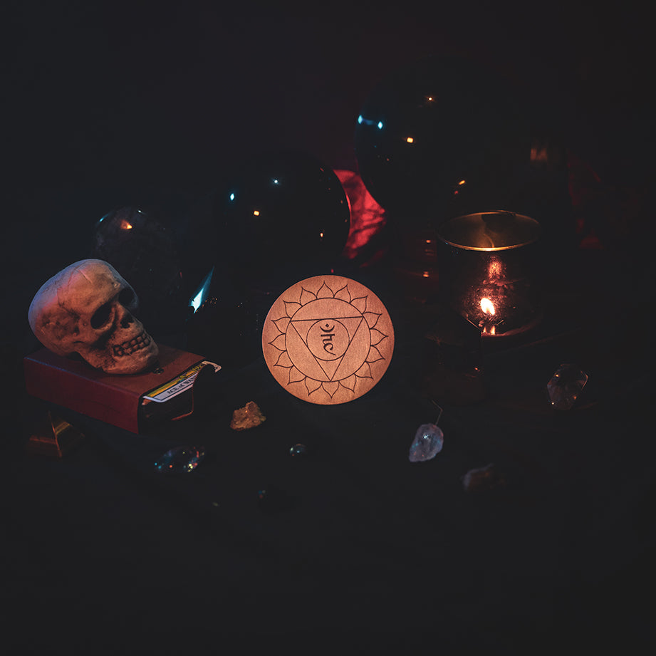 A circular wooden laser-cut crystal grid engraved with the throat chakra symbol. The grid sits amongst candles, crystal balls, a skull, tarot cards and assorted crystals.