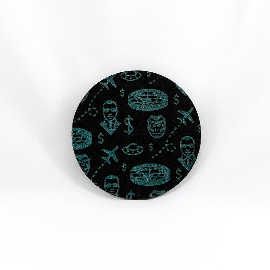 A round black coaster engraved with blue flying saucers, airplanes, a flat earth, dollar signs, a reptilian face, and a man with sunglasses and a black suit on. The coaster is on a white background.