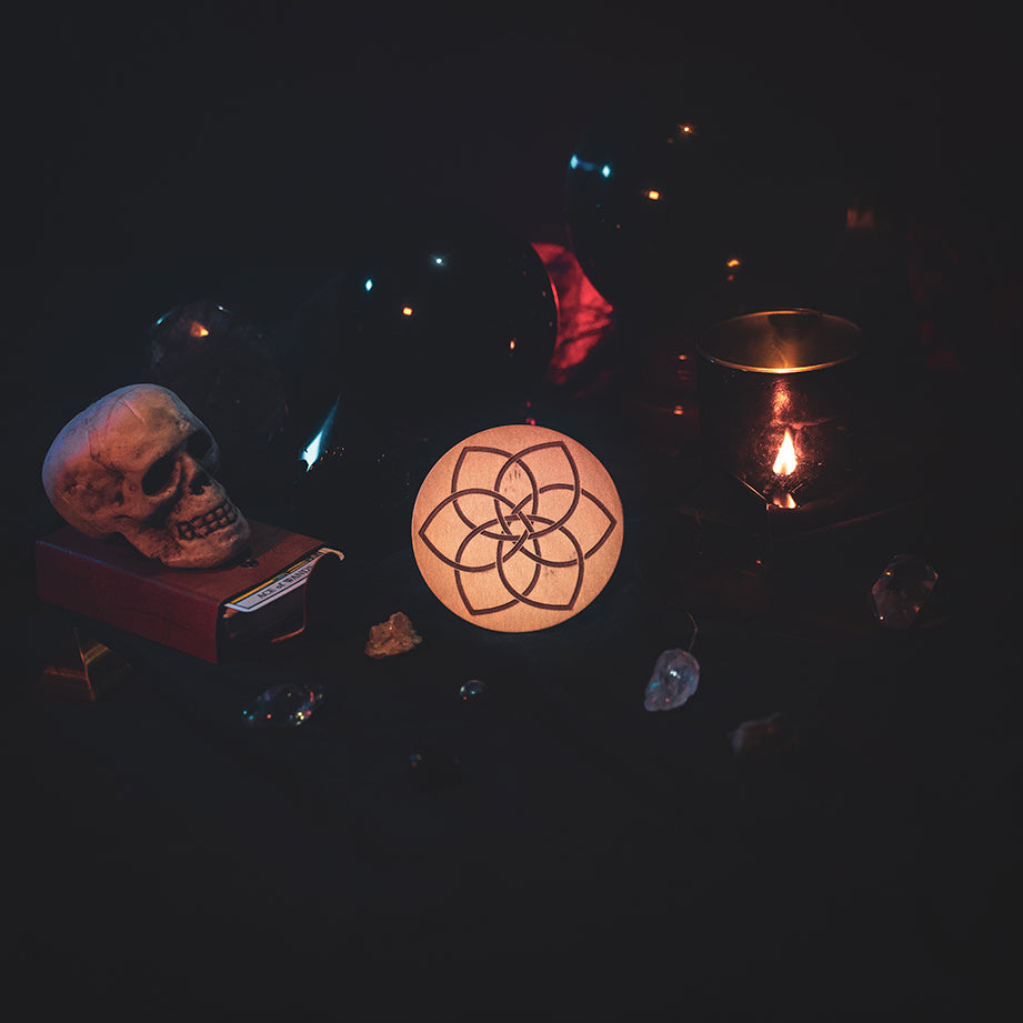 A circular wooden laser-cut crystal grid engraved with a Celtic flower knot. The grid sits amongst candles, crystal balls, a skull, tarot cards and assorted crystals.