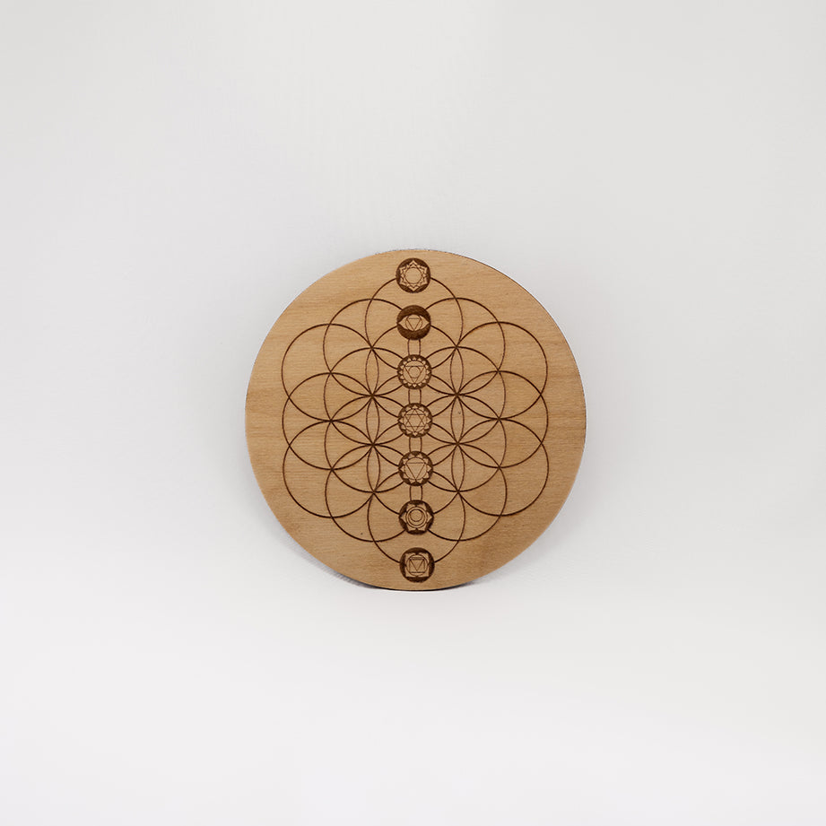 A circular wooden laser-cut crystal grid engraved with the Flower of Life and the seven Chakras with a plain white background. 