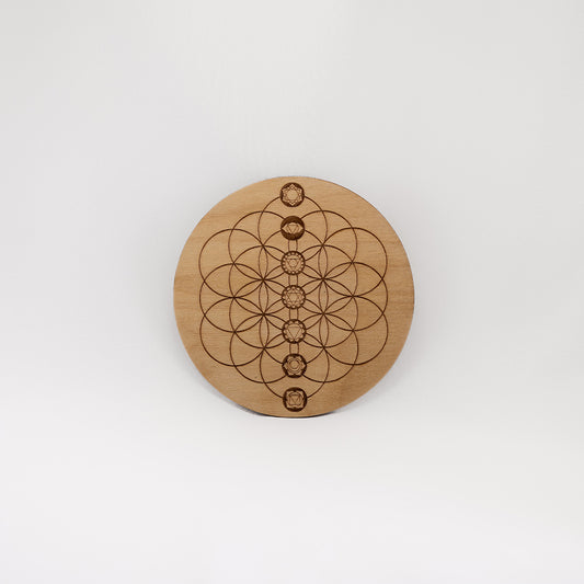 A circular wooden laser-cut crystal grid engraved with the Flower of Life and the seven Chakras with a plain white background.