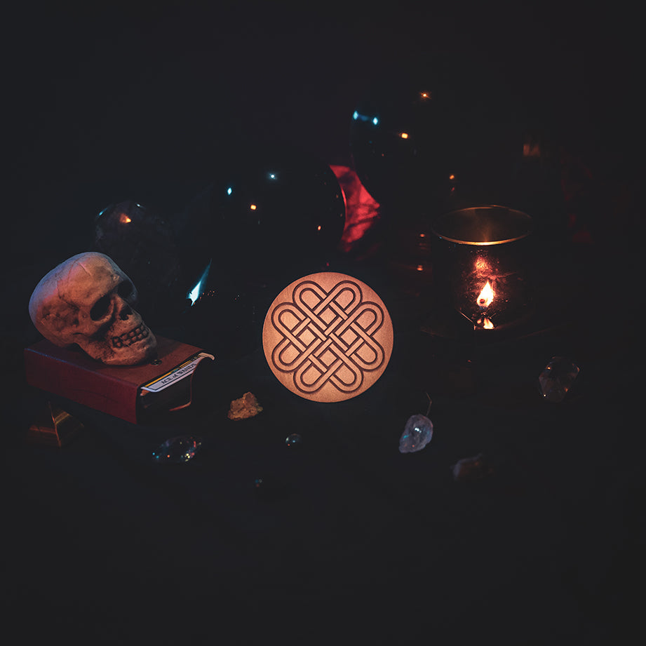 A circular wooden laser-cut crystal grid engraved with a Celtic knot. The grid sits amongst candles, crystal balls, a skull, tarot cards and assorted crystals.