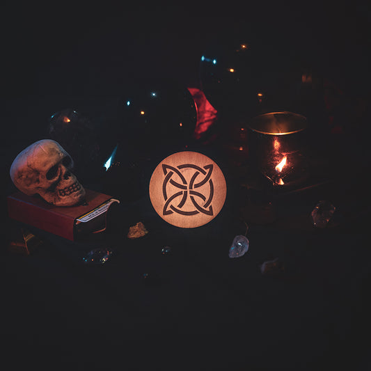 A circular wooden laser-cut crystal grid engraved with a Celtic knot. The grid sits amongst candles, crystal balls, a skull, tarot cards and assorted crystals.