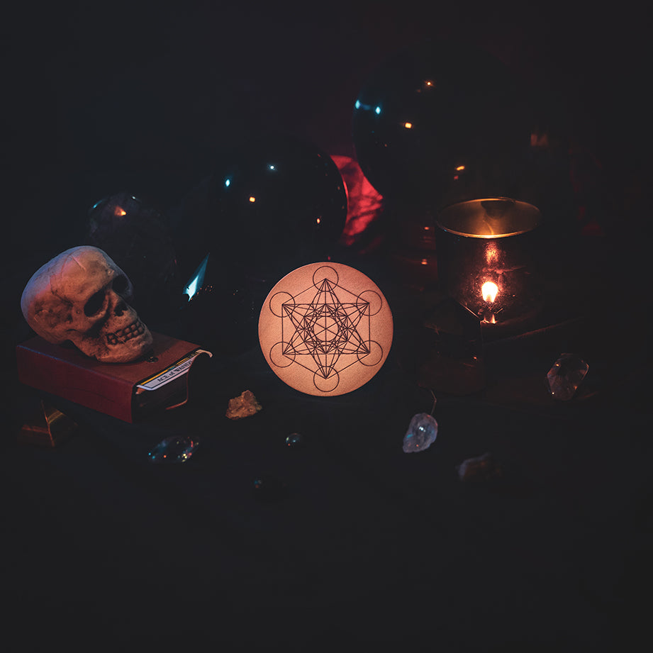 A circular wooden laser-cut crystal grid engraved with Metatron's Cube. The grid sits amongst candles, crystal balls, a skull, tarot cards and assorted crystals.