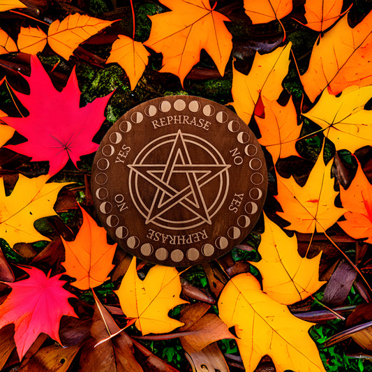 Circular Baltic birch laser-cut pendulum board engraved with the words "yes", "no", "rephrase",  a decorative moon phase border, and a large pentacle in the middle lays on the leaf covered ground. 