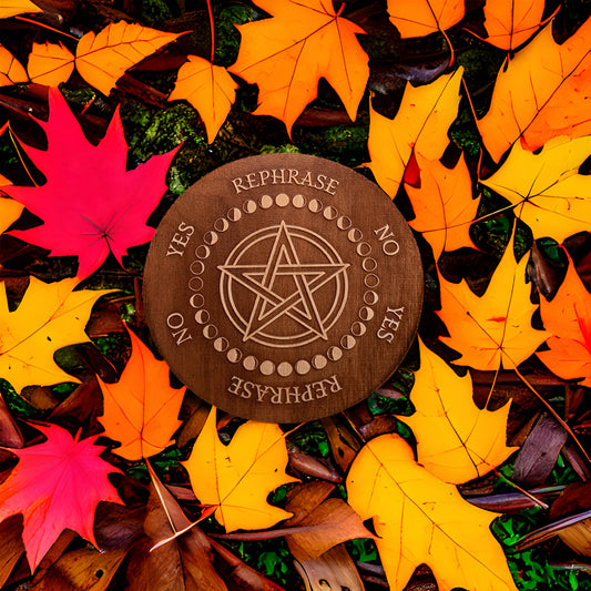 Circular Baltic birch laser-cut pendulum board engraved around its border with the words "yes", "no", "rephrase", the board has a pentacle in the centre encircled with the different moon phases and lays on the leaf covered ground.