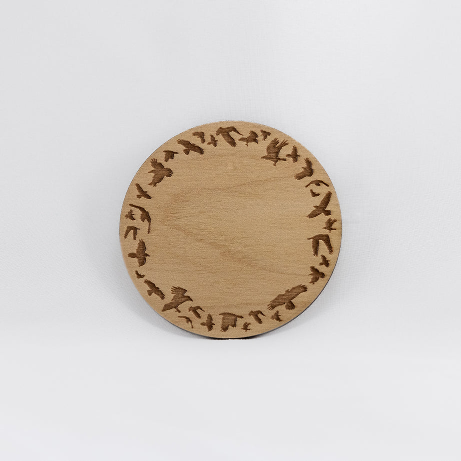 A circular wooden laser-cut crystal grid engraved with a border made up of ravens in flight with a plain white background. 